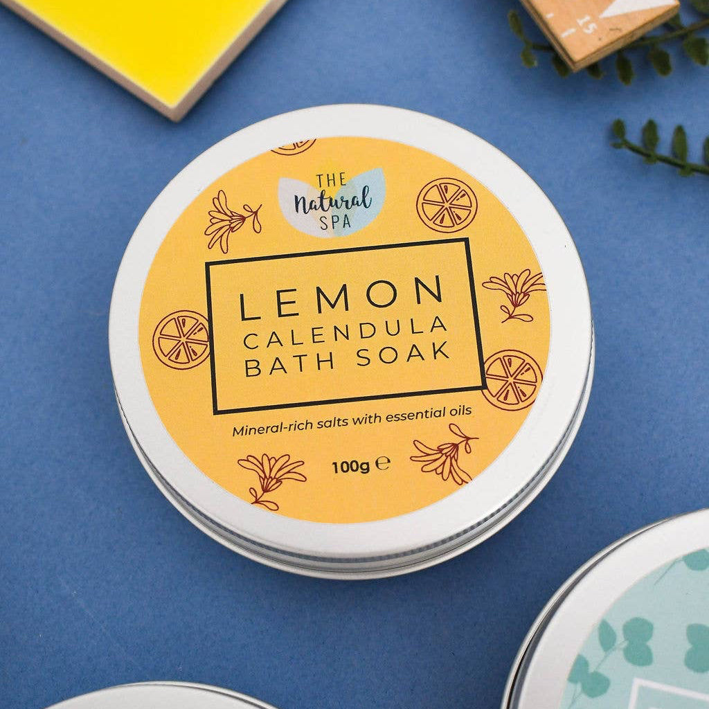 A round metal tin of The Natural Spa Lemon Calendula Bath Soak on a blue surface, with other bath soak tins partially visible around it and a yellow and wood block in the background.