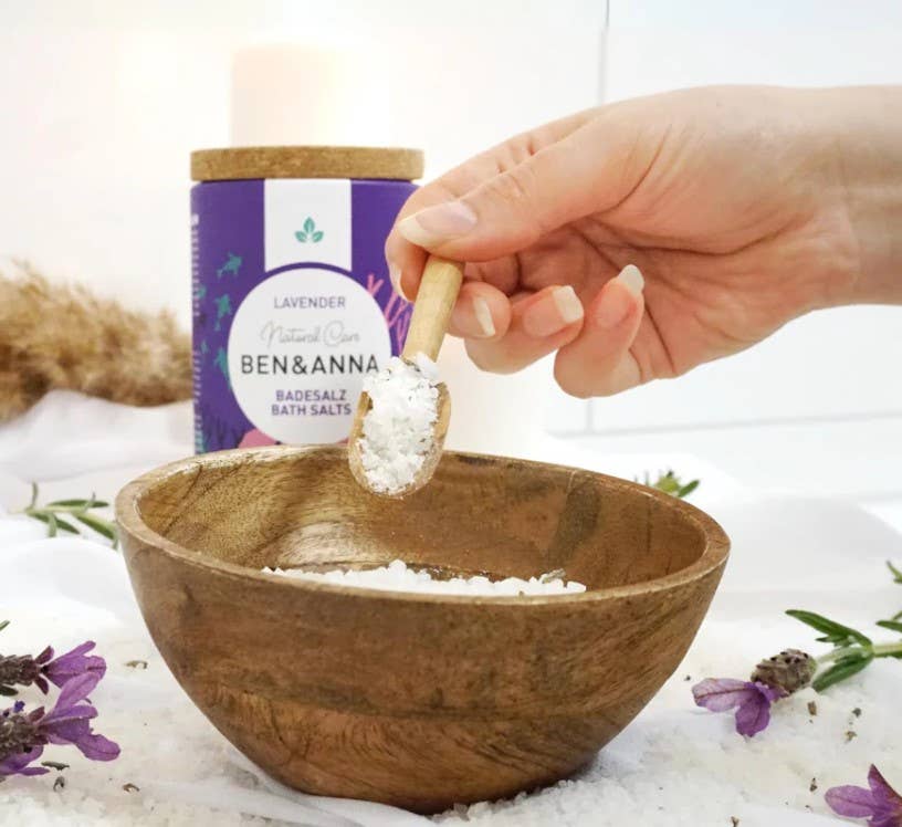 Person adding Ben & Anna bath salts to a wooden bowl with a the packaging of the product in the background.