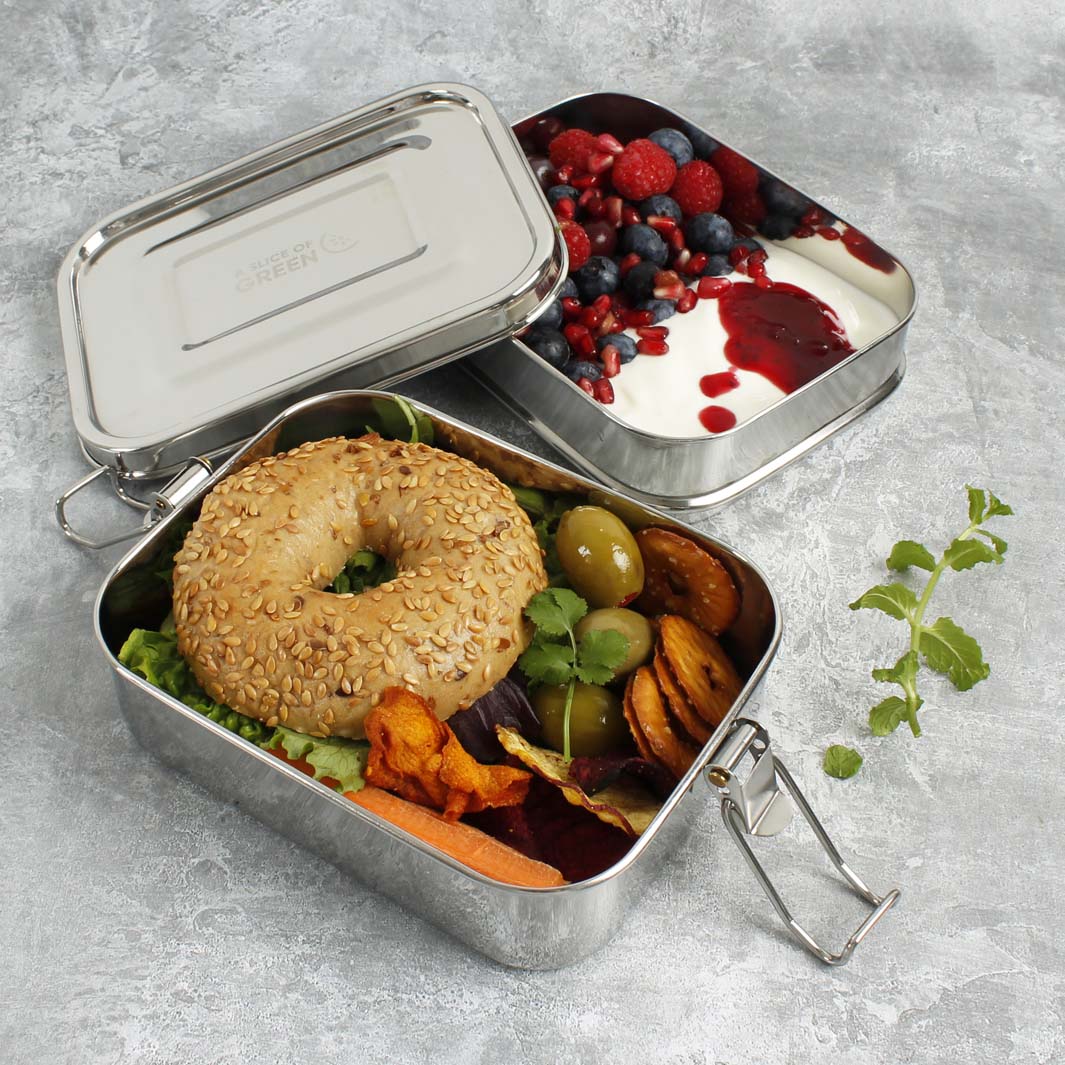 Two stainless steel lunch boxes filled with food on a light gray textured surface. One box is open and shows a seeded bagel, greens, carrots, and olives.
