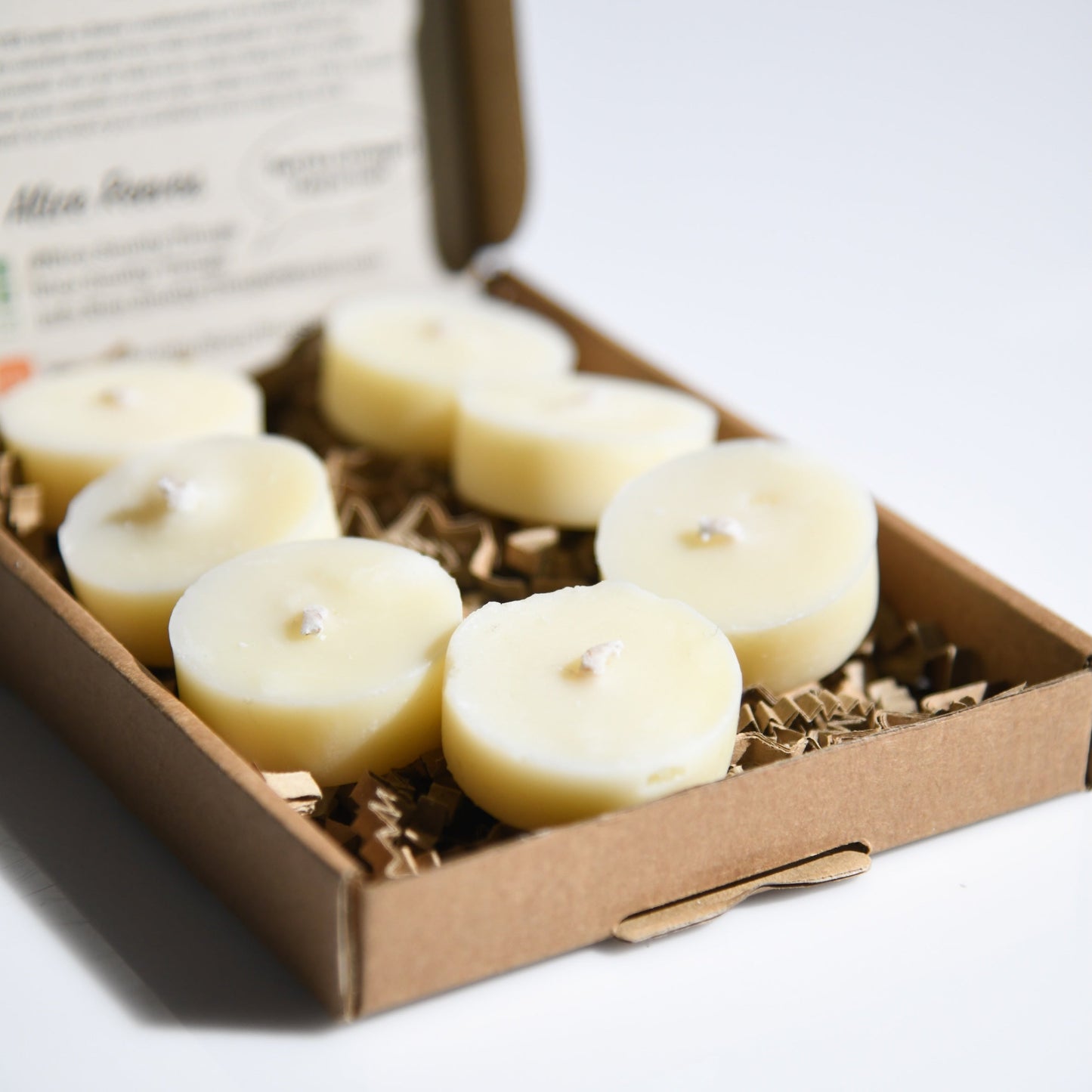 An open brown cardboard gift box containing six small round cream-colored tealight candles resting on shredded paper filler.