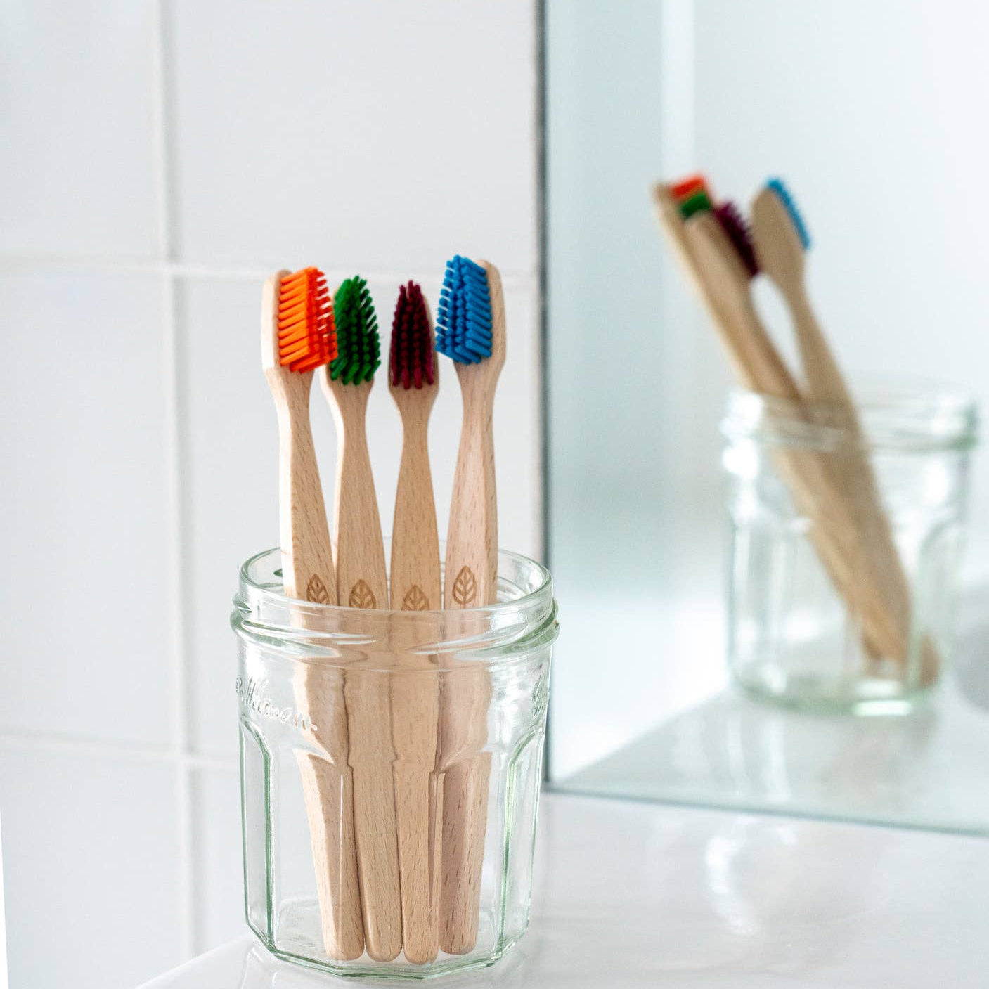 Four bamboo toothbrushes with colorful bristles in orange, green, brown, and blue, standing in a clear glass jar on a white bathroom counter with a mirror reflection.