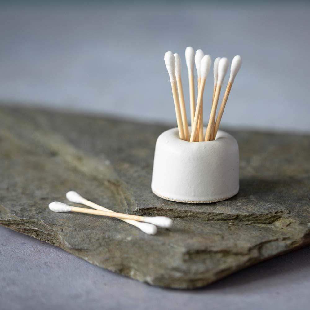 A small matte white cylindrical ceramic holder filled with wooden cotton swabs, placed on a dark stone slab against a soft gray background.