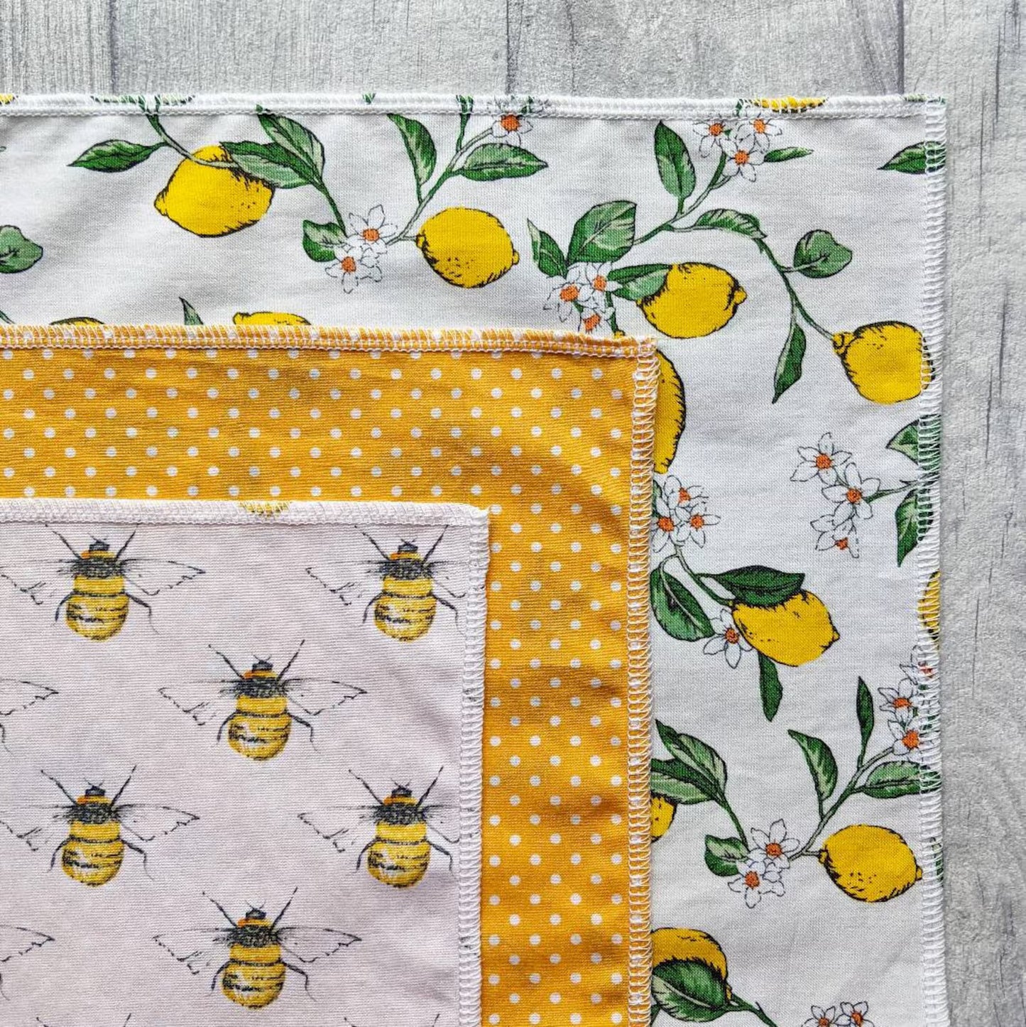 An overlay of three folded cotton fabric handkerchiefs on a gray wood-textured surface. The top fabric is white with yellow bumblebees and gray wings; the middle fabric is mustard yellow with white polka dots; the bottom fabric is off-white with green leaves and small white flowers.