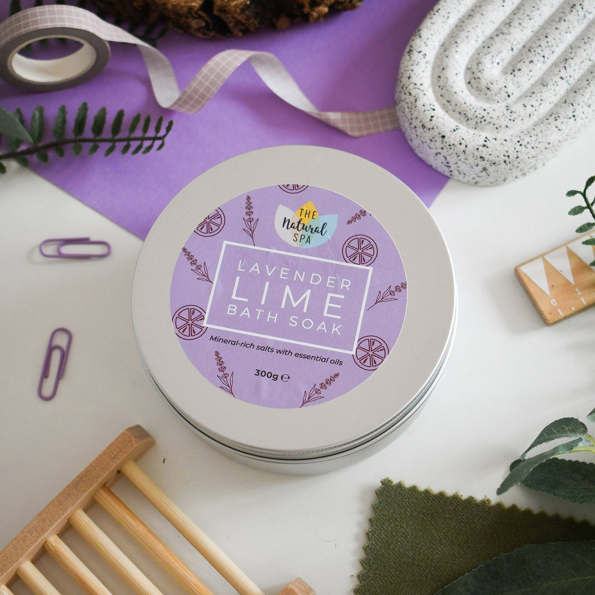 A round metal tin of Lavender Lime Bath Soak from The Natural Spa, styled in a flat-lay arrangement with purple paper, green leaves, and decorative paper clips on a white surface.
