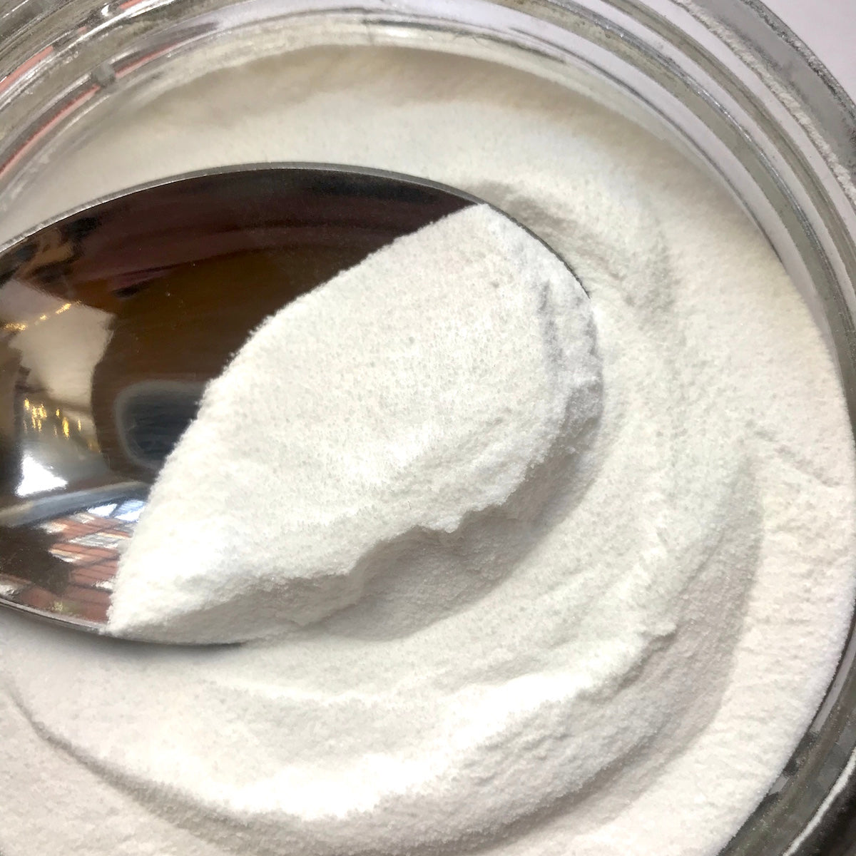 A close-up of a clear jar filled with white powder, with a metal spoon holding a scoop of the powder.