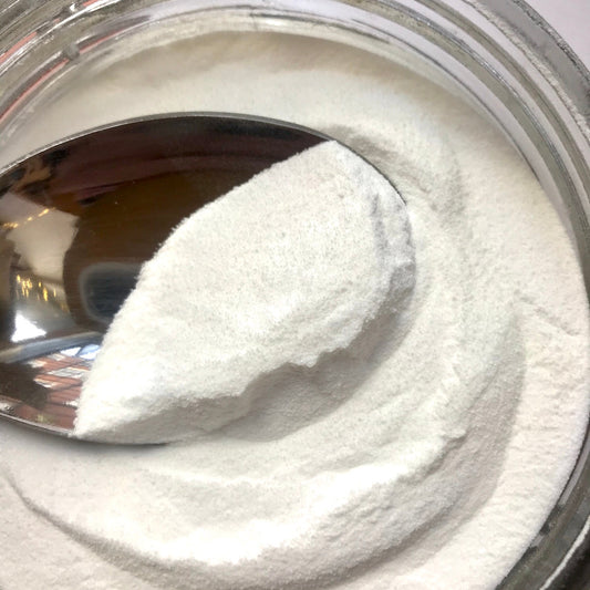 A close-up of a clear jar filled with white powder, with a metal spoon holding a scoop of the powder.