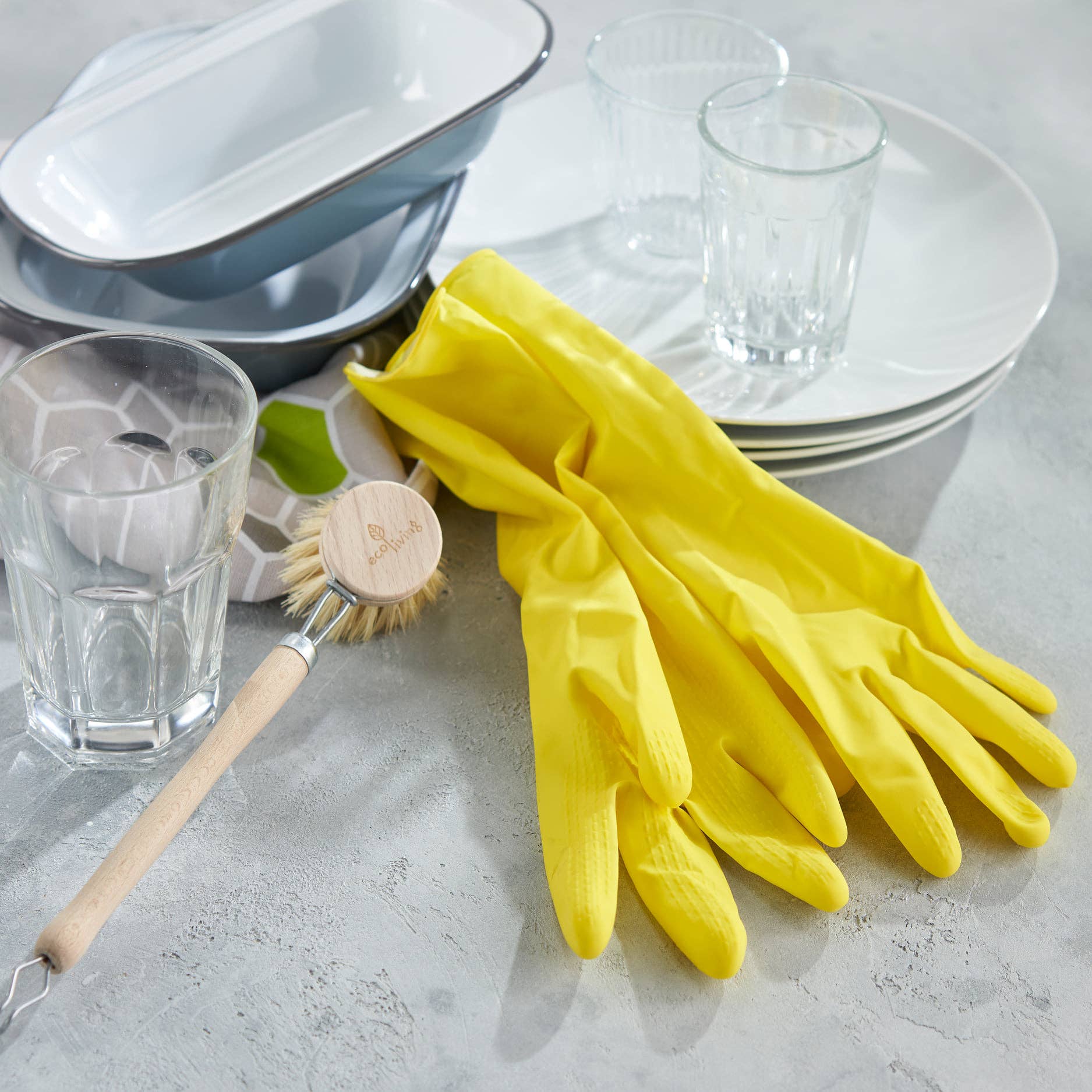 Yellow natural rubber cleaning gloves on a surface with glasses, a dish, and a scrubber.