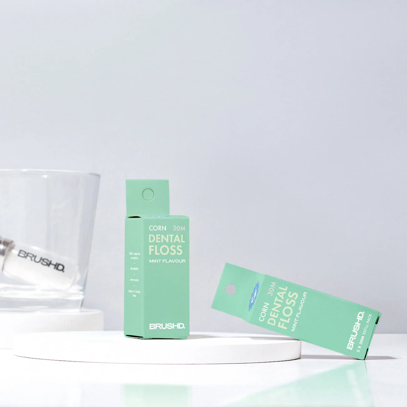 Brushd cornstarch dental floss in a green box with a glass and a glass floss dispenser in the background