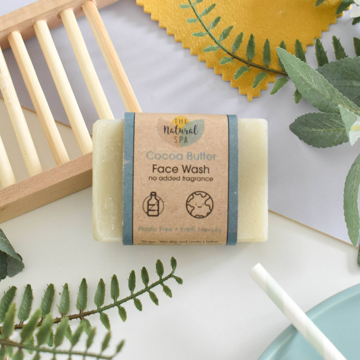Cocoa butter face wash bar with packaging on a white surface with green leaves and a wooden stand.