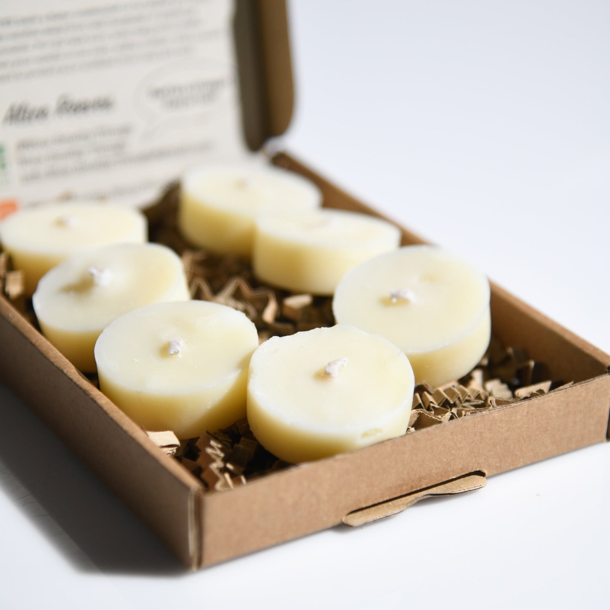 An open brown cardboard gift box containing six small round cream-colored tealight candles resting on shredded paper filler.