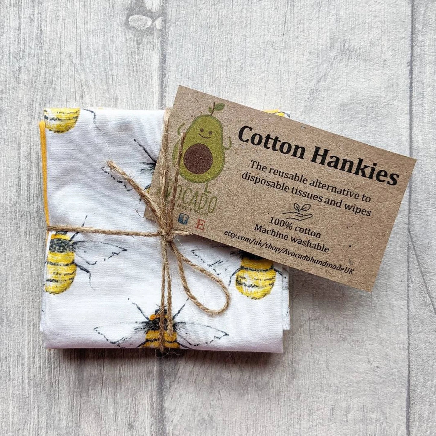 A folded set of reusable cotton hankies with a yellow and black bee print, tied with twine and featuring a brown paper tag that reads 'Cotton Hankies' with an avocado illustration, placed on a light gray wood background.
