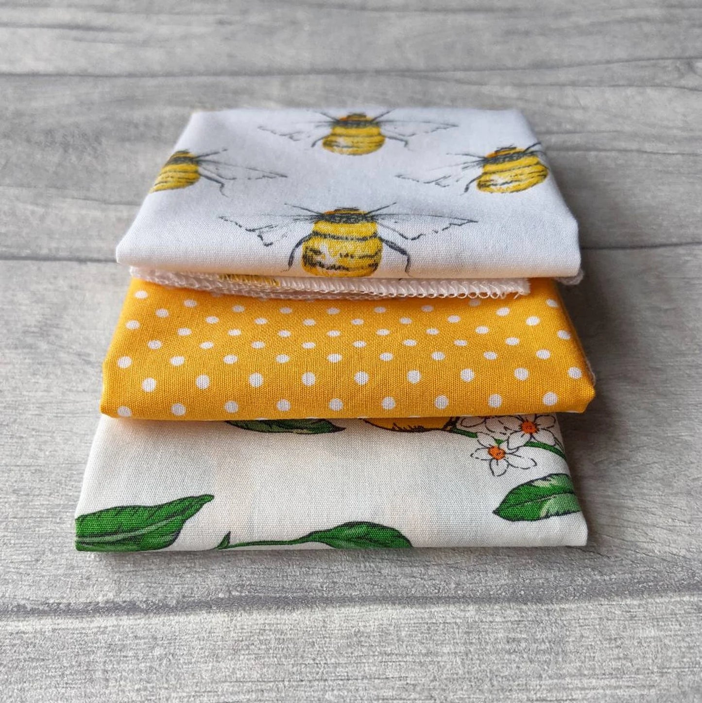A stack of three folded cotton fabric handkerchiefs on a gray wood-textured surface. The top fabric is white with yellow bumblebees and gray wings; the middle fabric is mustard yellow with white polka dots; the bottom fabric is off-white with green leaves and small white flowers.