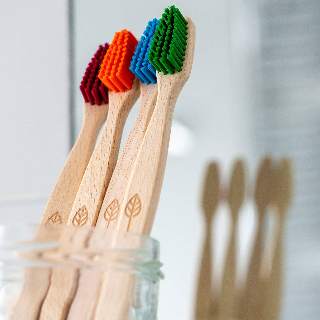 Four plant-based beechwood toothbrushes with colorful bristles in red, orange, blue, and green, standing in a clear glass jar. The toothbrush handles are light natural wood with a leaf engraving, set against a softly blurred bathroom background.