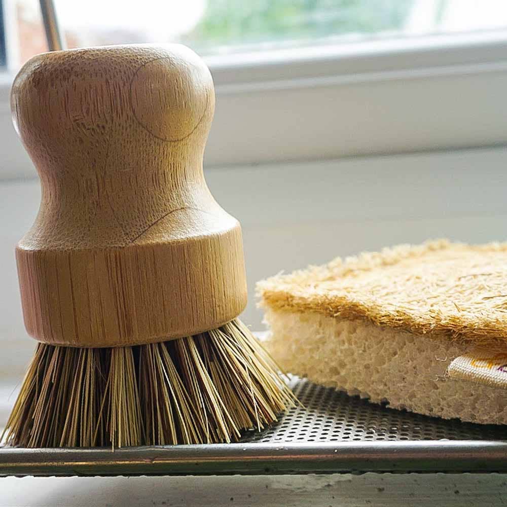 A wooden-handled dish brush with natural bristles sits on a metal tray next to a sponge in front of a window.