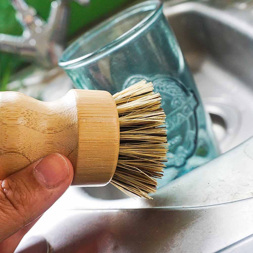 A hand holding a bamboo-handled dish brush with natural bristles, positioned over a stainless steel kitchen sink with a blue-tinted glass in the background.