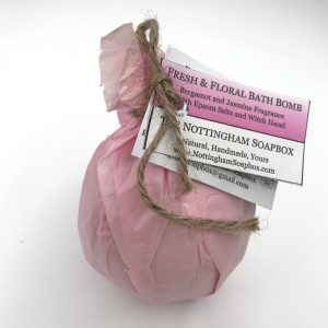 A wrapped bath bomb in a pink tissue paper package tied with twine and attached a paper label reading 'Fresh & Floral Bath Bomb' and 'NOTTINGHAM SOAPBOX', photographed on a plain white background.