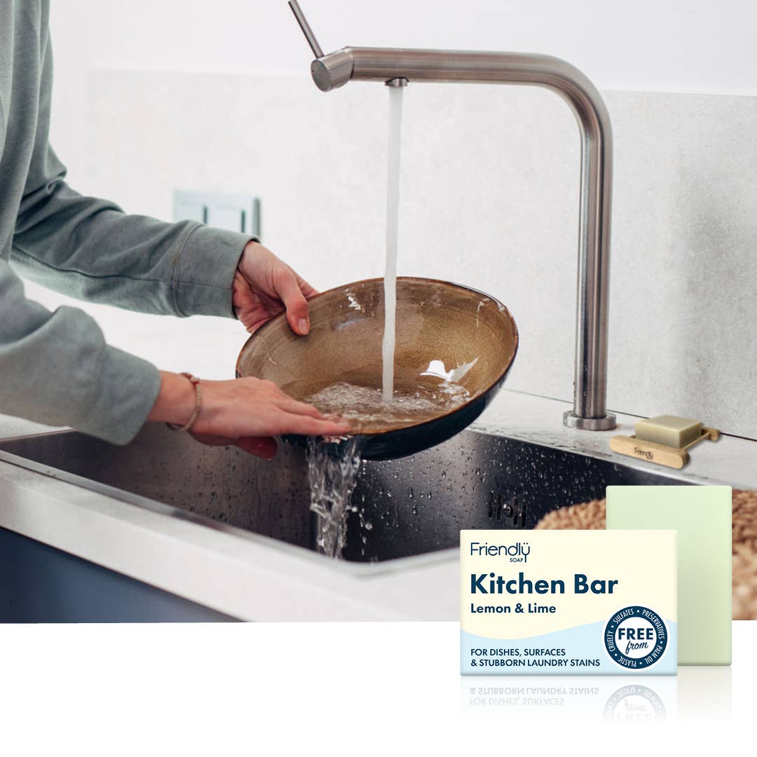 Person washing a bowl under running water in a kitchen sink with a Friendly Soap Kitchen Bar Lemon & Lime product displayed.