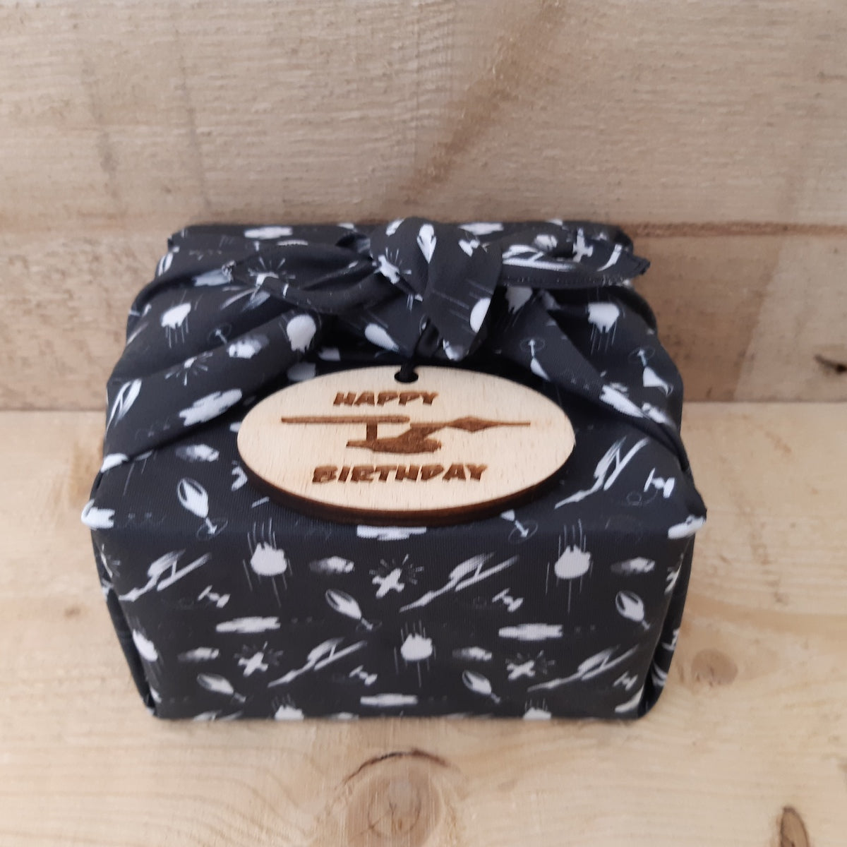 A square gift box wrapped in black and white Star Wars themed patterned fabric Furoshiki wrapping tied in a bow and a wooden oval tag that reads 'HAPPY BIRTHDAY'.