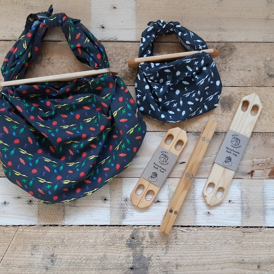 Two black patterned fabric Furoshiki wraps made into bags by use of wooden Patchin handles, sitting alongside 3 different sized pairs of Patchin handles arranged on a rustic light wood plank background, one Patching handle set showing the magnets in the handle which snap the bags shut