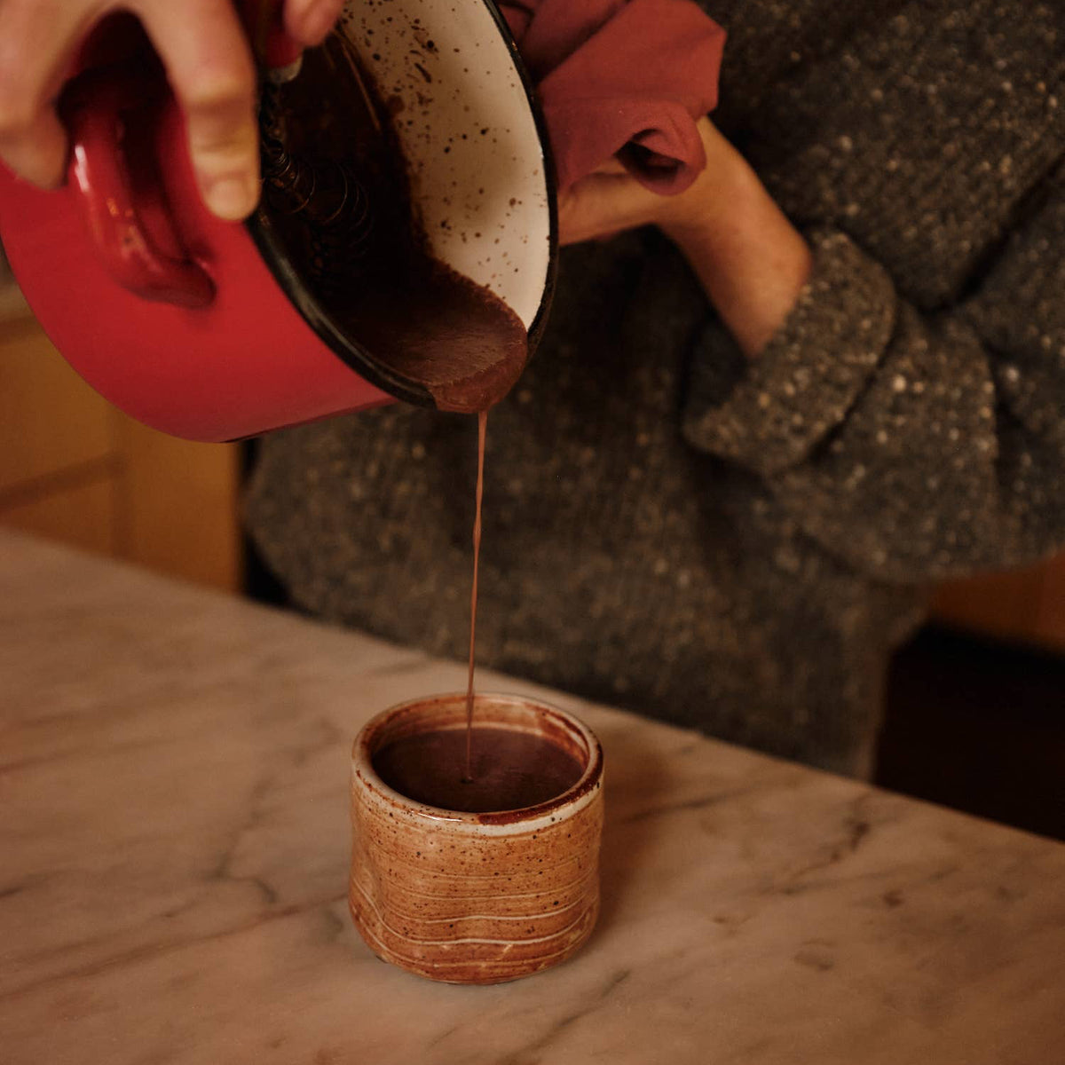 Person pouring a dark hot chocolate liquid from a red saucepan  into a small brown cup on a marble surface.