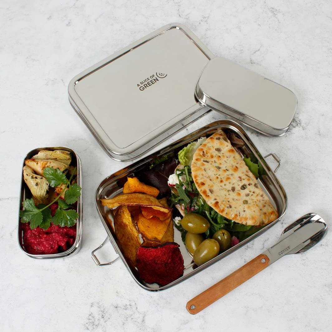 A stainless steel lunch box set on a light marble surface, shown open with a salad wrap, roasted vegetables, olives, and dips. The set includes two rectangular bento-style boxes, a smaller rectangular container, and a wooden-handled metal utensil.