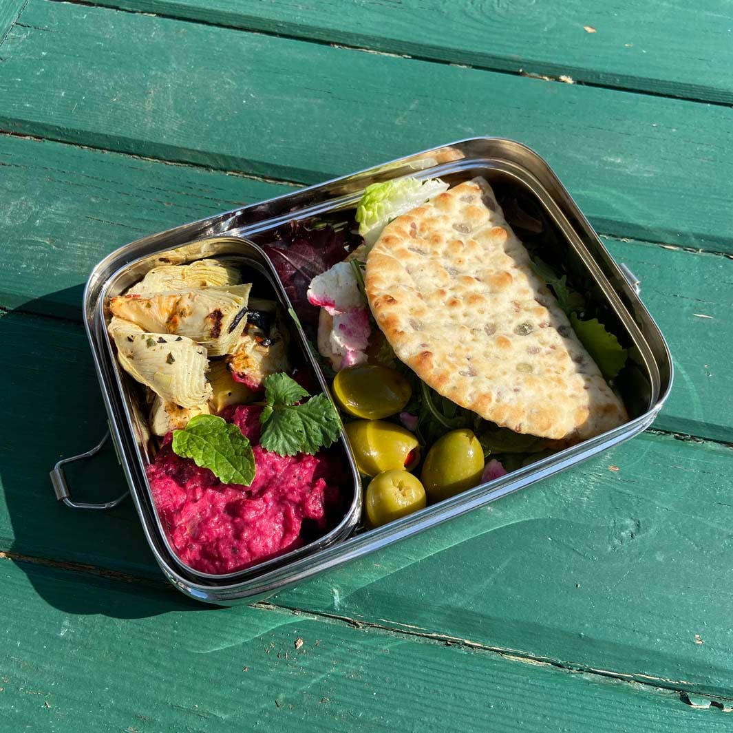 A rectangular metal lunch box with an added insert box  filled with a Mediterranean-style meal, placed on a green wooden table. The meal includes a folded flatbread, a pink dip, green olives, and a wrap with mint.