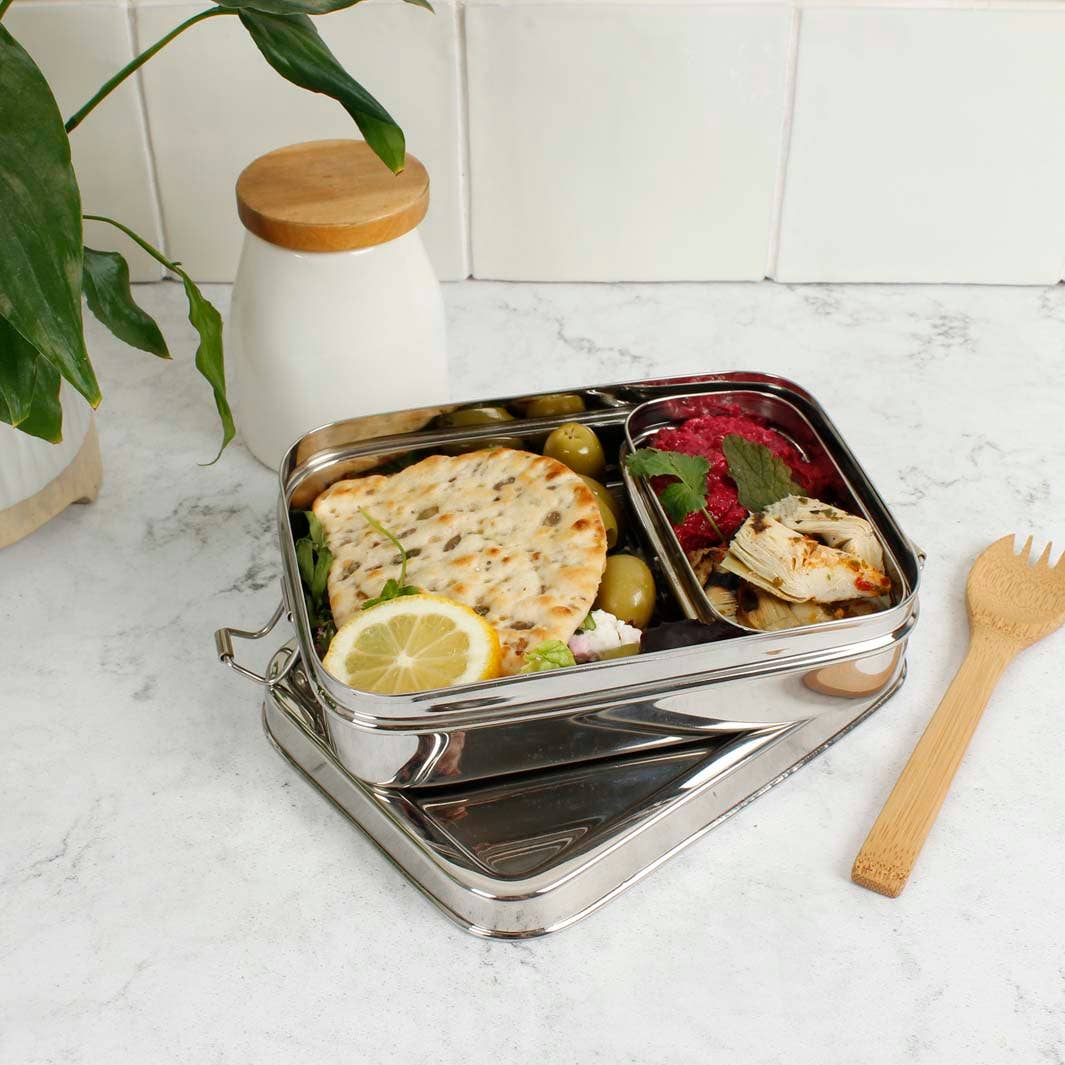 A stainless steel compartment lunch box with a lid, shown filled with food on a light marble surface. The box has an added insert boxcontaining flatbread, olives, a lemon slice, and a red salad, with a wooden utensil beside it and a white jar with a wooden lid in the background.