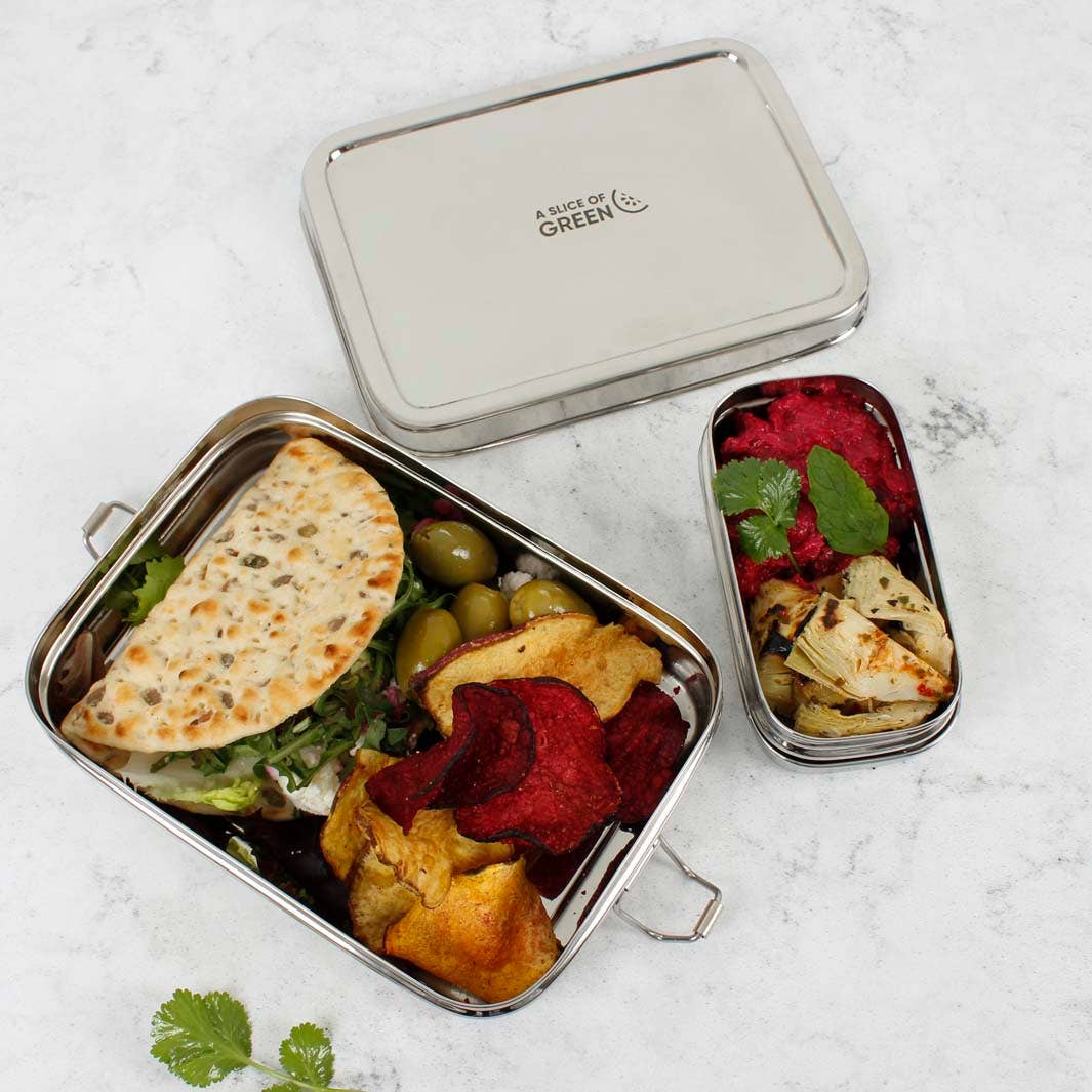 A stainless steel lunch box with lid on a light marble surface, filled with a sandwich, olives, chips, and a side of roasted vegetables. Alongside a smaller insert container 