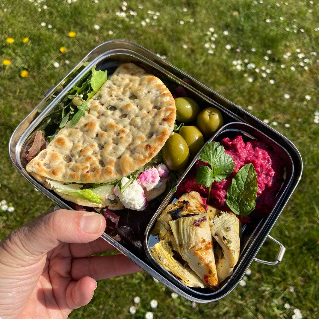 Hand holding a metal lunch box with food against a grassy background