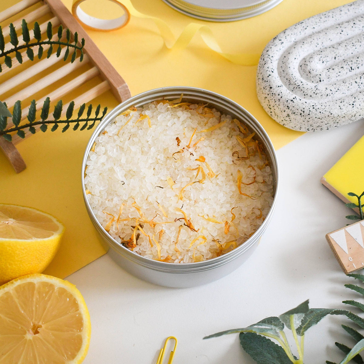 Metal tin filled with lemon bath salts on a yellow background with lemons and calendula  flowers.