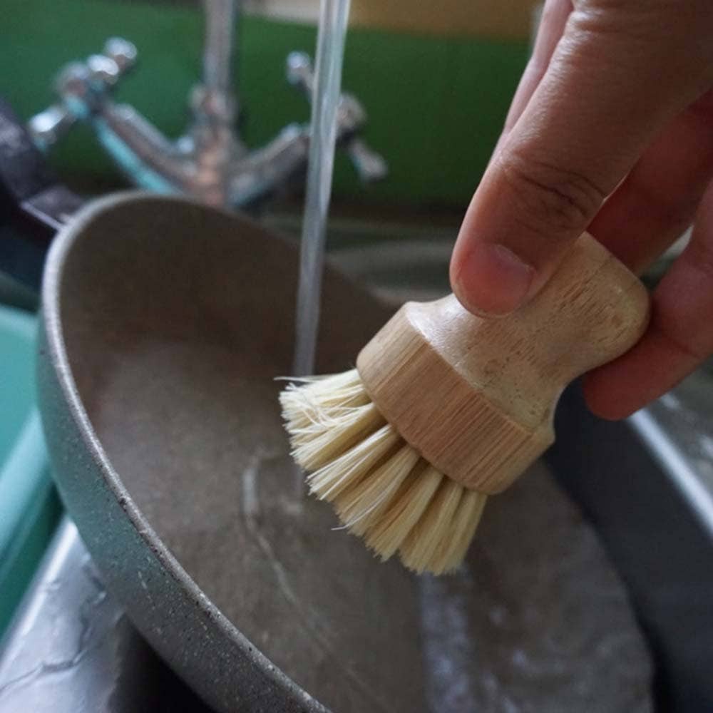 A hand holding a natural bamboo dish brush with soft beige bristles under running water in a sink.