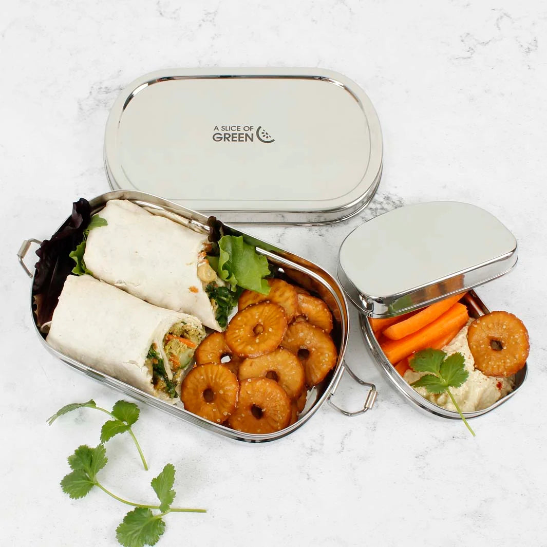 A stainless steel bento lunch box set on a light marble surface, shown open with wraps, fruit slices, crackers, and carrot sticks arranged inside. The lid is placed behind and a smaller inset view shows the contents in detail.