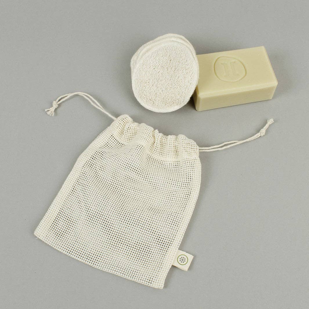 A natural cotton mesh bag with drawstrings, shown alongside a washable make up pads and a bar of soap on a neutral gray background.