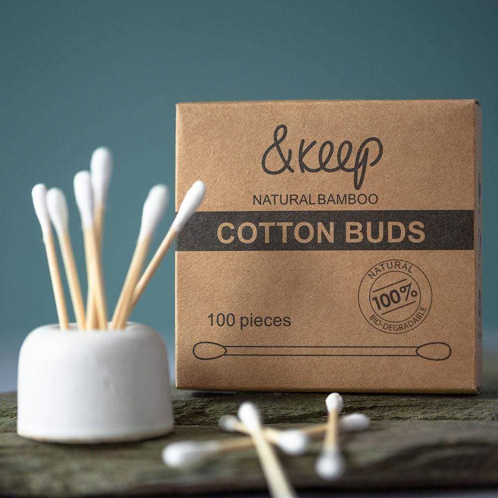 A kraft card box of natural bamboo cotton buds, next to a white container holder some of the cotton buds, and some shown in front of the box, all on a wooden surface.