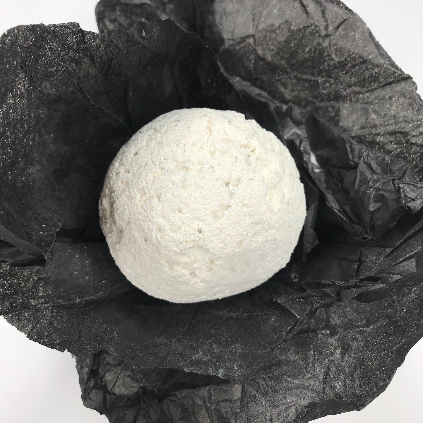 A round white bath bomb resting on crumpled black tissue paper against a plain white background.