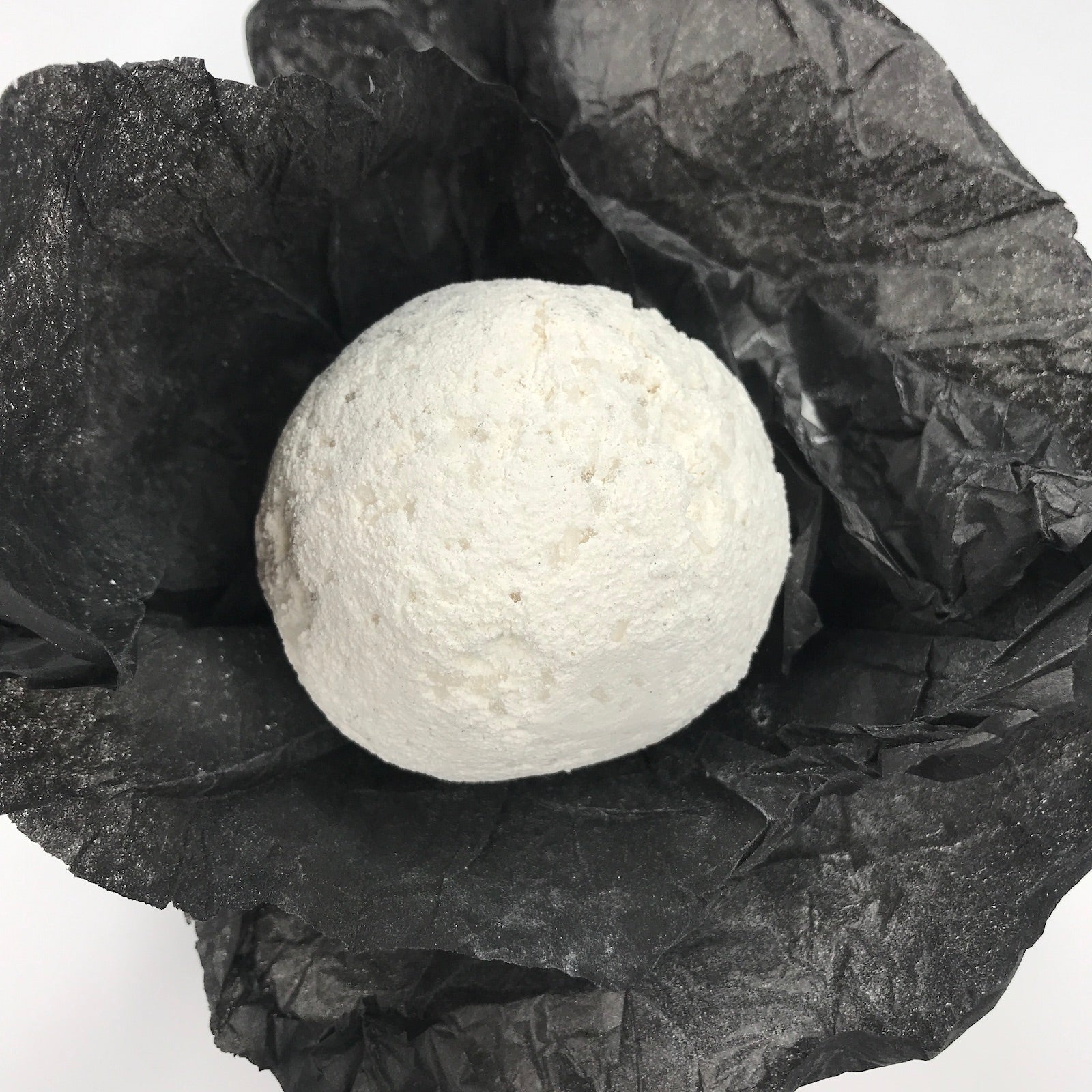 A round white bath bomb resting on crumpled black tissue paper against a plain white background.