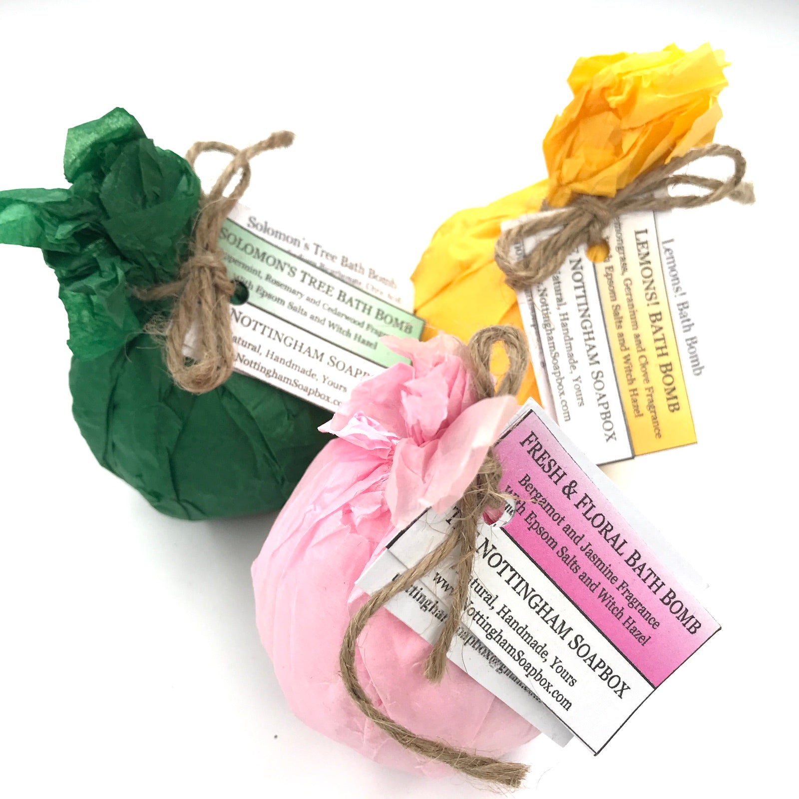Three packaged bath bombs in green, pink, and yellow tissue paper bags tied with twine, each with a paper label from Nottingham Soapbox on a white background.
