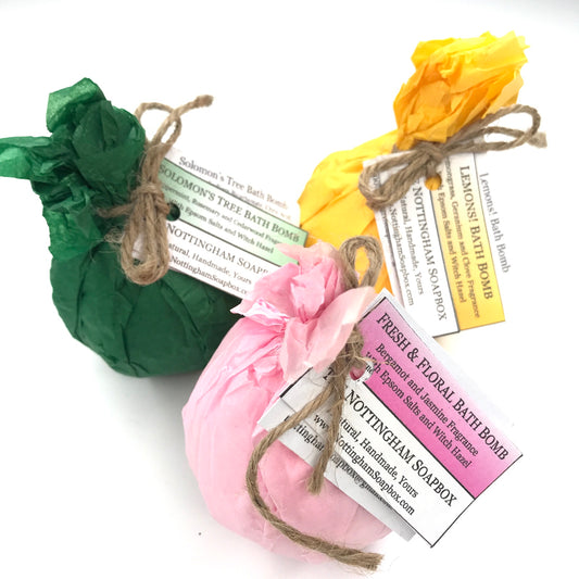 Three packaged bath bombs in green, pink, and yellow tissue paper bags tied with twine, each with a paper label from Nottingham Soapbox on a white background.