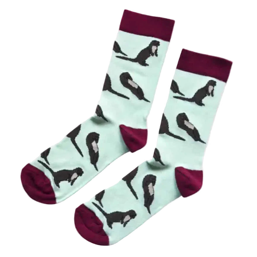 A pair of crew socks with a mint green background, featuring a black and white  otter pattern and deep burgundy cuffs, heels, and toes, displayed against a solid pale pink background.