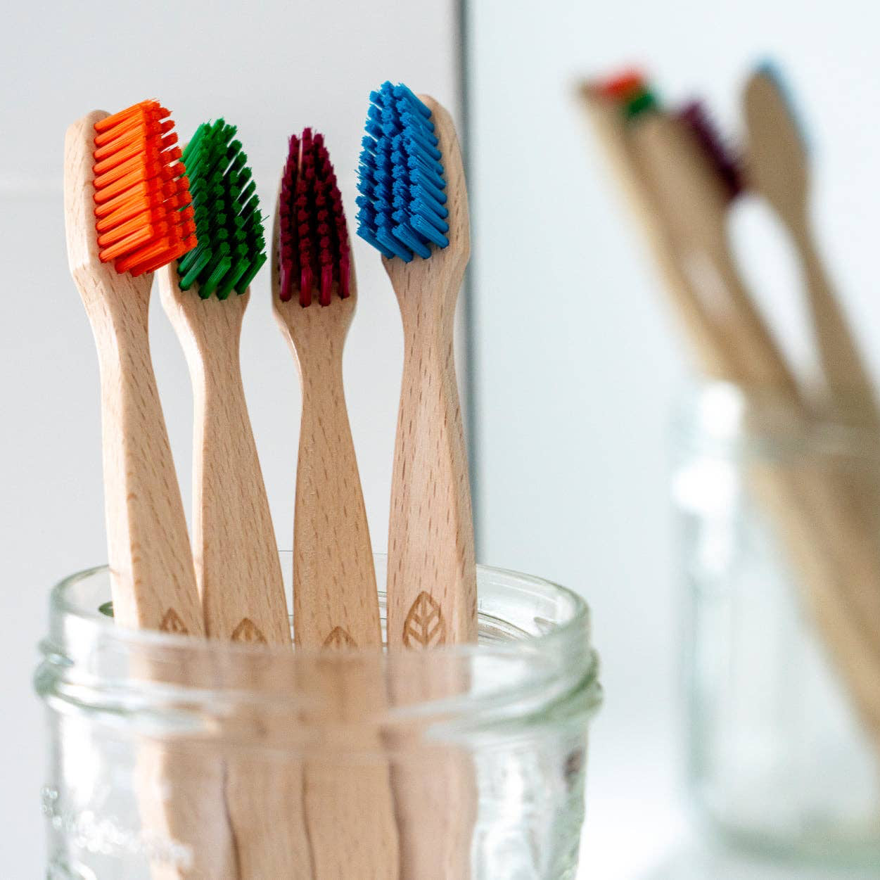 Four beechwood toothbrushes with colorful bristles (orange, green, dark red, and blue) standing in a clear glass jar, with a blurred second jar in the background.
