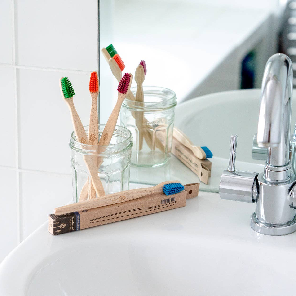 A close-up of a white bathroom sink with chrome taps, showing a glass toothbrush holder and several bamboo toothbrushes in clear jars, with a mirror reflecting the scene.