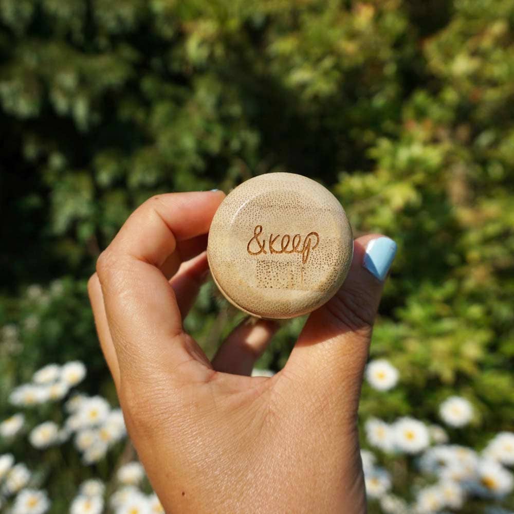 A hand holding a round, light beige bamboo pot brush handle with the brand name '&keep' printed on top, set against a blurred background of green foliage and white daisies.