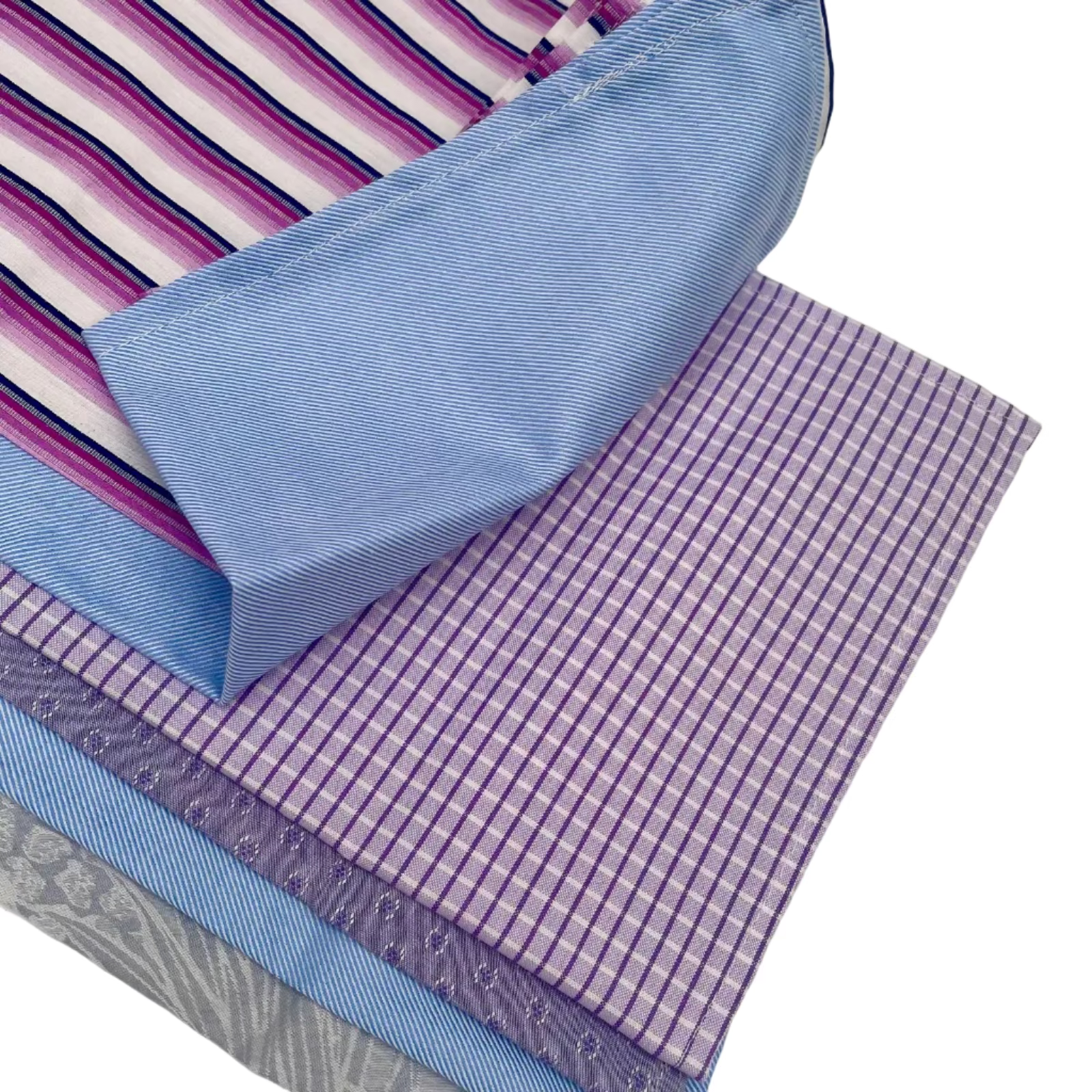 A close-up of folded and layered cotton handmade handkerchiefs in purple, pink, blue, and gray patterns, including stripes, gingham, and damask textures, arranged on a white surface.