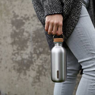 Person holding a silver water bottle with a wooden cap set against a textured wall.
