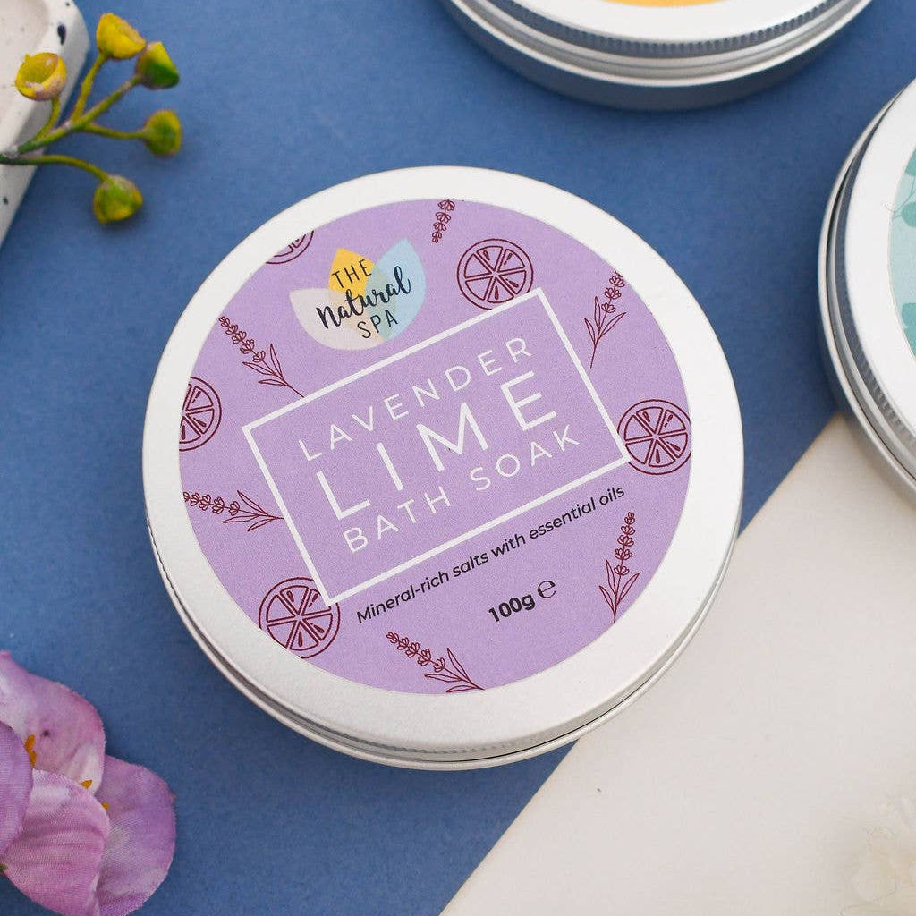 A round tin of lavender lime bath soak from The Natural Spa, featuring a purple and white label, styled on a blue background with other bath soaks and decorative flowers.