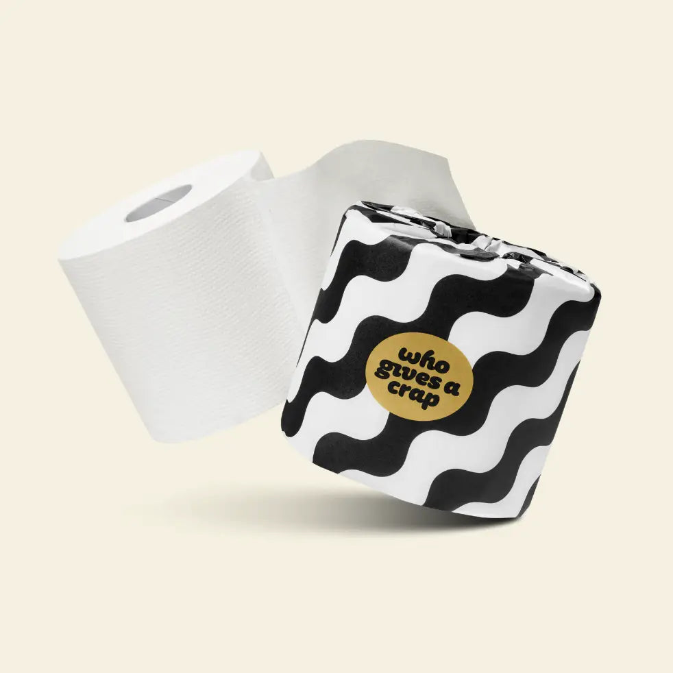 A roll of white toilet paper next to a square tissue pack with a black and white wavy pattern and a gold circular sticker that reads 'who gives a crap' in black text, set against a light beige background.