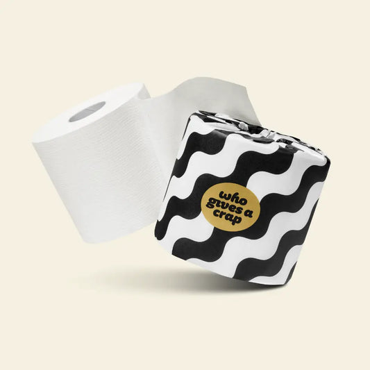 A roll of white toilet paper next to a square tissue pack with a black and white wavy pattern and a gold circular sticker that reads 'who gives a crap' in black text, set against a light beige background.