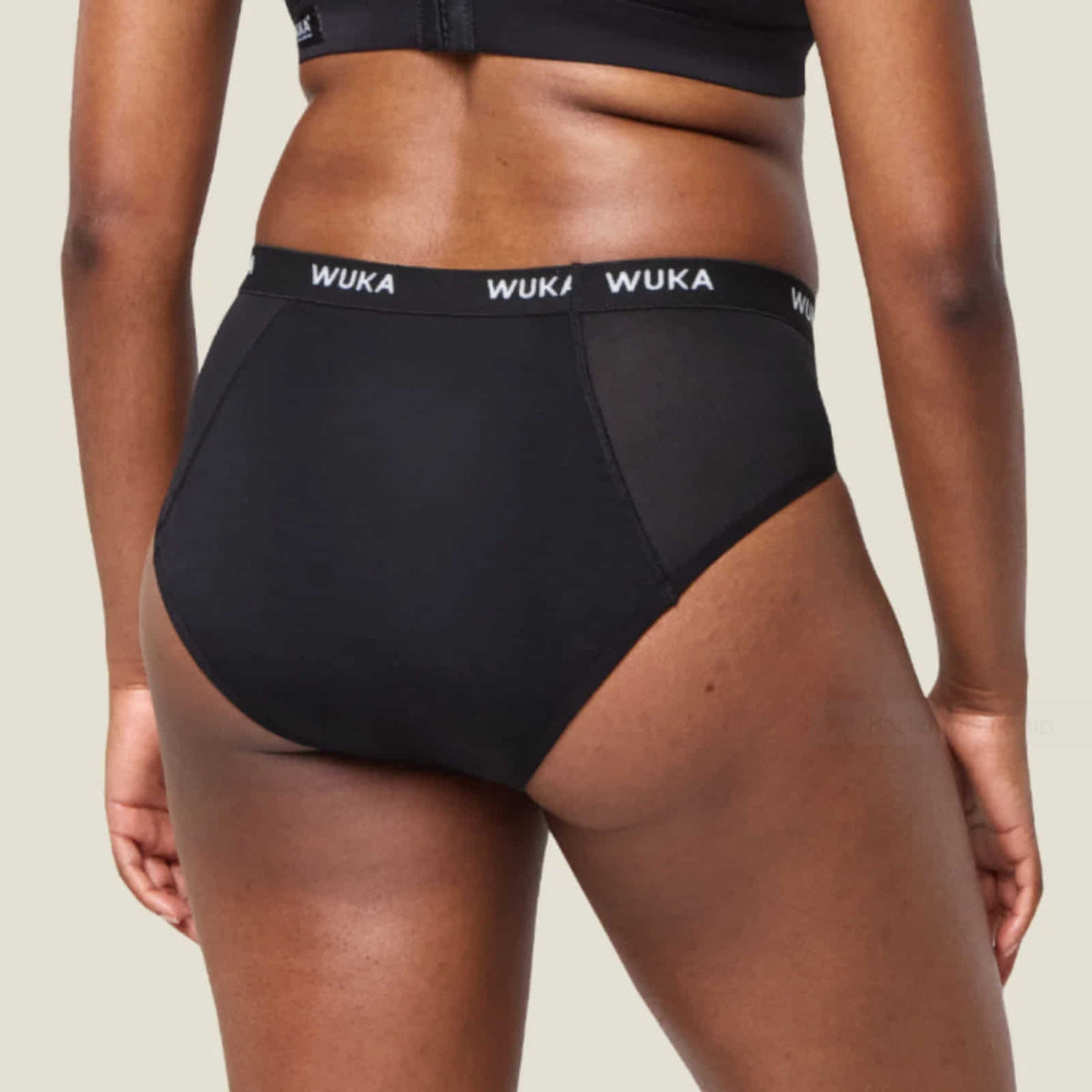 A close-up of a model from the back wearing black high-waisted briefs with a black waistband featuring the white WUKA brand name repeated across it.